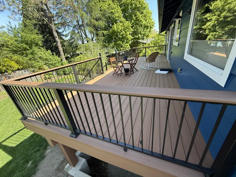 Raised brown PVC deck with black aluminum railing, a wood-toned top rail and a table and chairs beside a blue home