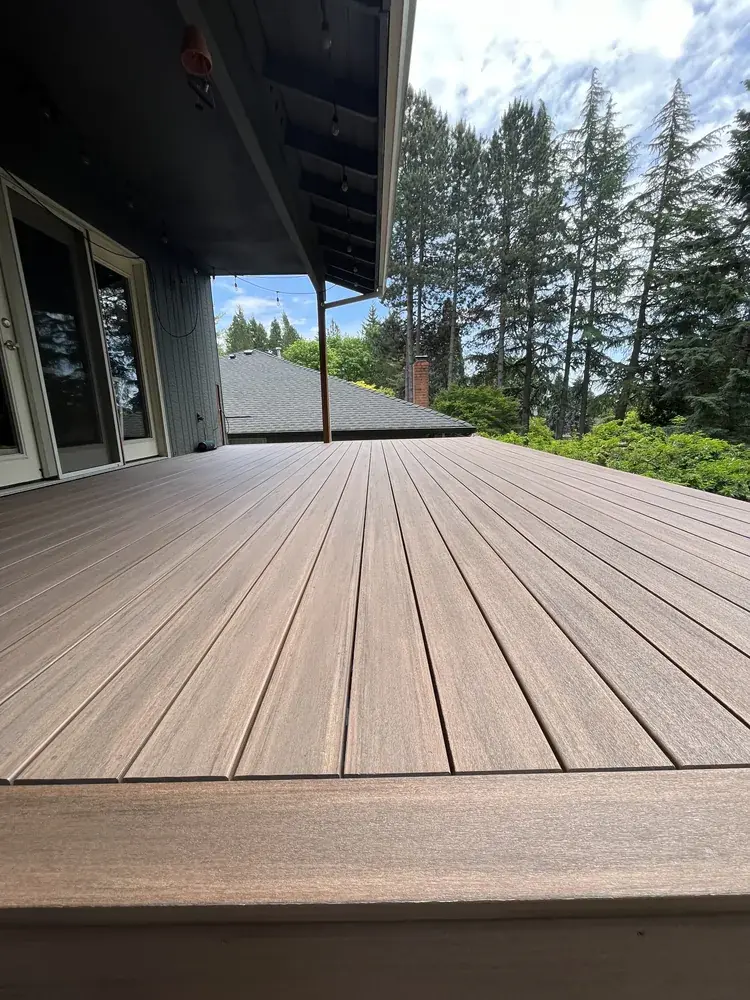 Low view along a brown PVC deck under a covered edge strung with lights, firs beyond the rooftop
