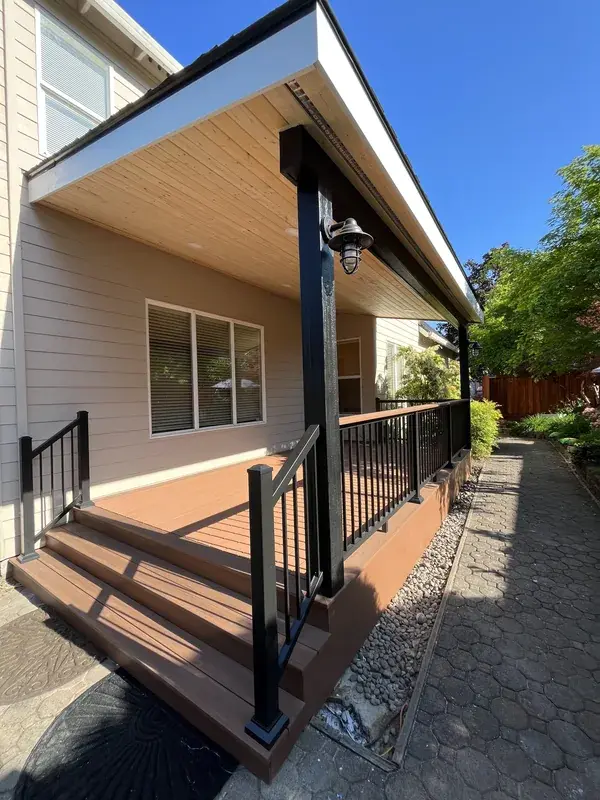 The same side of the house with a roofed deck built along it, black railing, steps down to the path and a light under the eave
