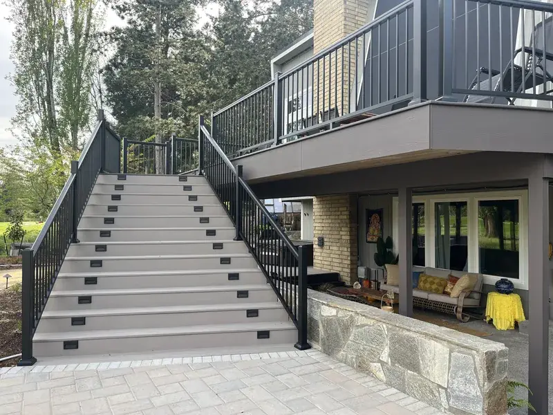 The same house with the finished deck above the patio, a wide flight of lit stairs coming down to the pavers and black aluminum railing throughout