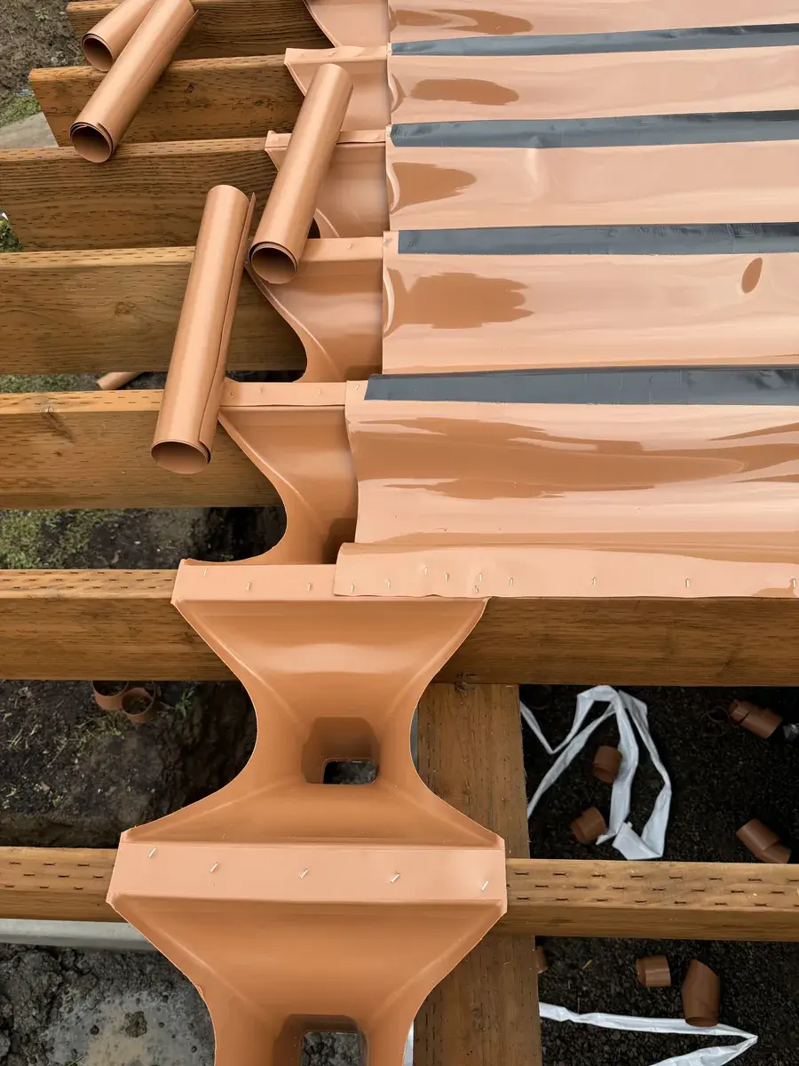 Drainage troughs and downspouts fitted between taped joists, carrying water off the frame before the deck boards go down