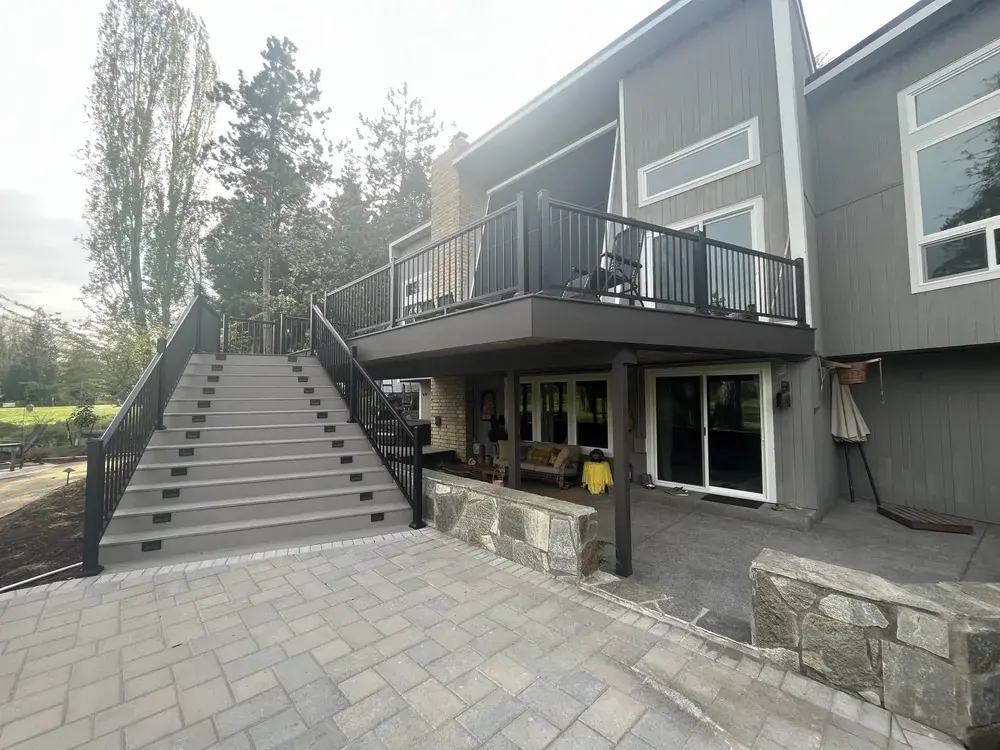 Elevated deck with black aluminum railing above a stone patio, a wide flight of lit stairs running down to the pavers