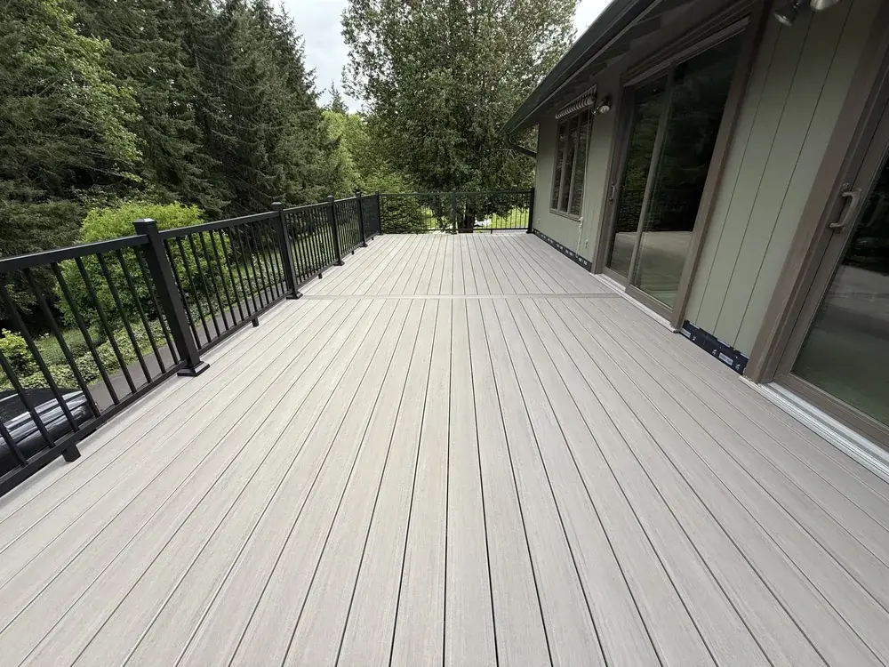 Long greige PVC deck running past sliding doors with black aluminum railing and firs along the far side